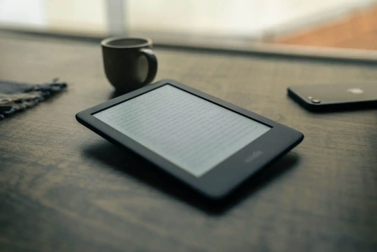 A Kindle kept on the table along with a phone and a black cup